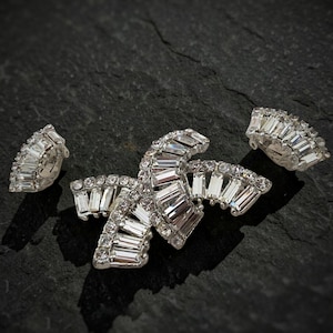 May include: A set of three silver-toned, rhinestone-encrusted jewellery pieces. The set includes a large, stylized butterfly-shaped brooch and two matching clip-on earrings. The rhinestones are clear and rectangular, set in a silver metal.