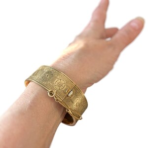 May include: A gold-colored hinged bracelet with intricate floral and geometric patterns. The bracelet is secured with a clasp and safety chain. The design features repeating circular motifs and detailed engravings, creating a vintage aesthetic.