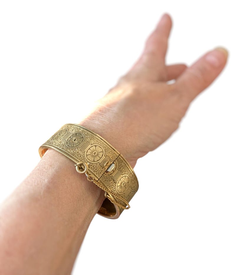May include: A gold-colored hinged bracelet with intricate floral and geometric patterns. The bracelet is secured with a clasp and safety chain. The design features repeating circular motifs and detailed engravings, creating a vintage aesthetic.