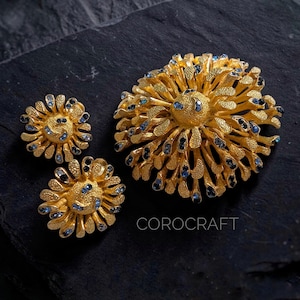 Peut inclure: Un ensemble de bijoux doré comprenant une grande broche et deux boucles d'oreilles assorties. Les pièces sont d'inspiration florale, avec des détails complexes et de petits accents bleus et noirs. Le mot "COROCRAFT" est imprimé en blanc.