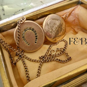 Puede incluir: Un collar dorado con un colgante redondo con un diseño de herradura con pequeñas gemas oscuras. El collar se muestra dentro de una caja dorada vintage con un segundo colgante ovalado. El texto "F&B" es visible.