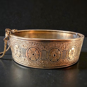 May include: A gold-toned bangle bracelet with an intricate floral design. The bracelet features a series of stylized flower motifs and decorative patterns. A small chain is attached to the clasp. The bracelet is a vintage piece.