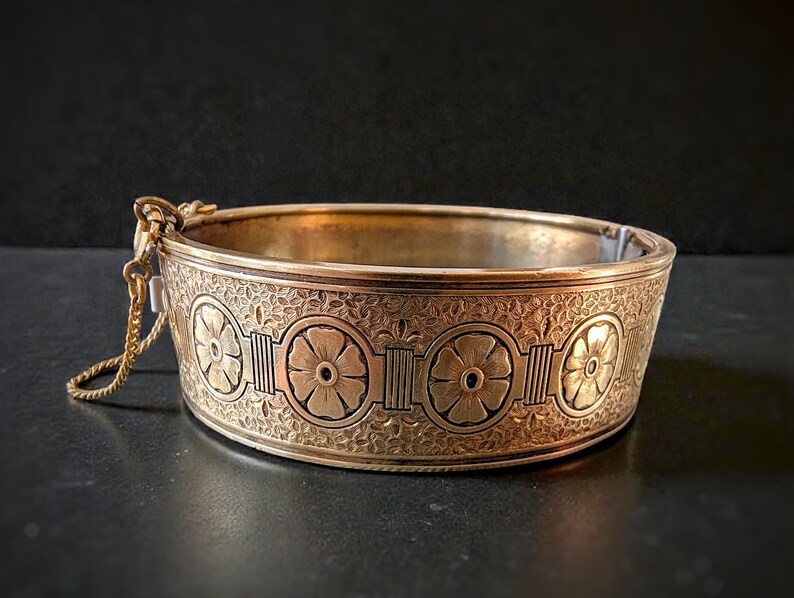 May include: A gold-toned bangle bracelet with an intricate floral design. The bracelet features a series of stylized flower motifs and decorative patterns. A small chain is attached to the clasp. The bracelet is a vintage piece.
