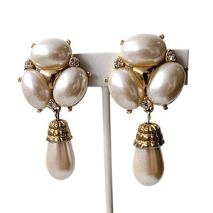 Puede incluir: Un par de pendientes vintage con grandes detalles de perlas de imitación. Cada pendiente tiene un grupo de tres perlas ovaladas, con una perla más pequeña y una gota dorada en forma de campana. Los brillantes transparentes añaden brillo.