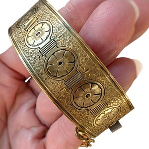 May include: A gold-toned hinged bangle bracelet. The bracelet features a repeating floral design within circular frames, connected by rectangular bars. The background is filled with a delicate, textured pattern. The clasp is visible on the side.