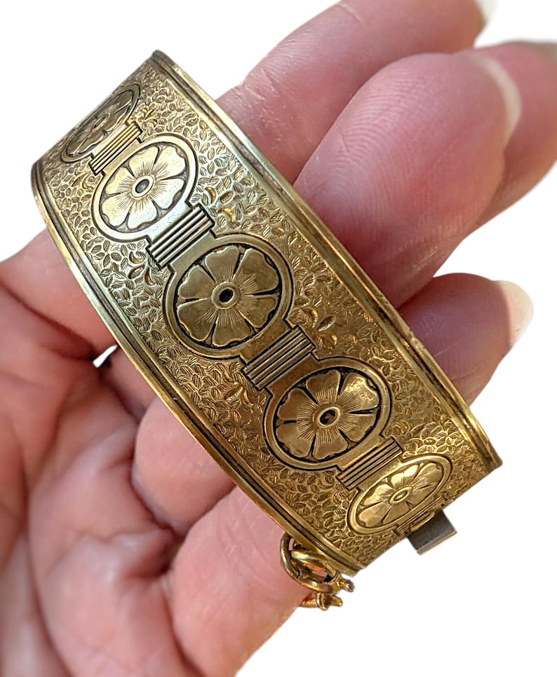 May include: A gold-toned hinged bangle bracelet. The bracelet features a repeating floral design within circular frames, connected by rectangular bars. The background is filled with a delicate, textured pattern. The clasp is visible on the side.