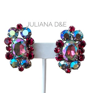 May include: A pair of vintage Juliana D&E clip-on earrings. Each earring features a large, iridescent oval stone surrounded by smaller, ruby-red and aurora borealis crystals. The earrings are displayed on a white stand.