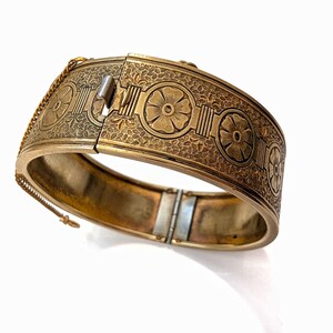 May include: A gold-toned hinged bangle bracelet with intricate floral and geometric patterns. The bracelet features a secure clasp and a safety chain. The design includes repeating flower motifs and detailed etched textures, creating a vintage aesthetic.