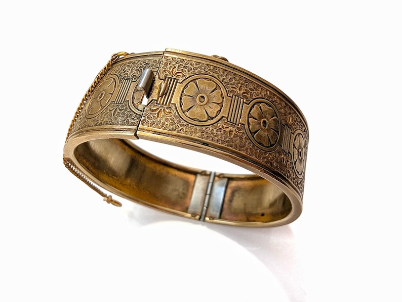 May include: A gold-toned hinged bangle bracelet with intricate floral and geometric patterns. The bracelet features a secure clasp and a safety chain. The design includes repeating flower motifs and detailed etched textures, creating a vintage aesthetic.