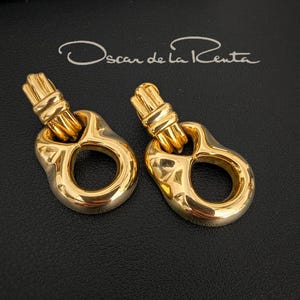 May include: A pair of gold-coloured earrings with a distinctive design. Each earring has a textured top section linked to a rounded, open-centre shape. The earrings are shown on a dark surface, with the designer's signature, "Oscar de la Renta," visible.