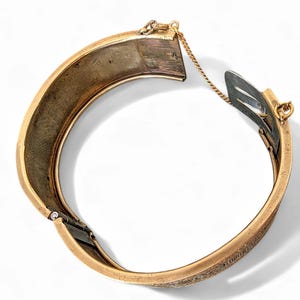 May include: A gold-toned hinged bangle bracelet with an engraved design. The bracelet is open, revealing the interior and clasp mechanism. A small gold chain connects the two halves.