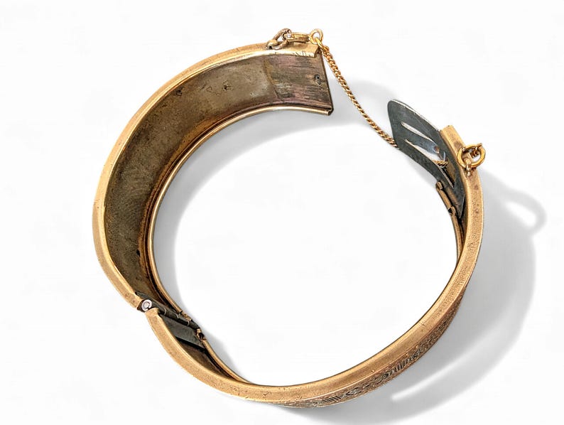 May include: A gold-toned hinged bangle bracelet with an engraved design. The bracelet is open, revealing the interior and clasp mechanism. A small gold chain connects the two halves.