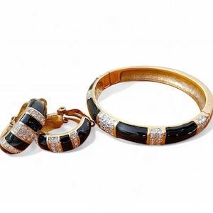 Può includere: Un set di gioielli color oro con un braccialetto e orecchini a cerchio abbinati. Il braccialetto e gli orecchini presentano un design a strisce in smalto nero con accenti di cristallo trasparente. I gioielli sono su sfondo bianco.