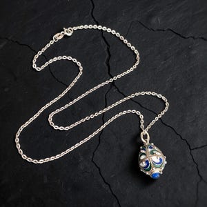 May include: A silver chain necklace featuring an egg-shaped pendant. The pendant is decorated with intricate silver filigree patterns and inlaid with blue and green enamel sections, finished with a small blue stone at the bottom point.