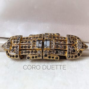May include: Vintage Coro Duette brooch with a gold-tone metal setting. The brooch features a geometric design with clear and black rhinestones. The text "CORO DUETTE" is displayed below the brooch.