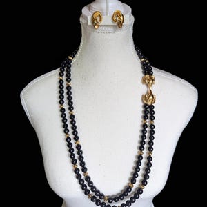 Puede incluir: Un collar de cuentas negras con un cierre de cabeza de carnero dorado y pendientes a juego de cabeza de carnero dorado. El collar es de doble cadena y las cuentas son redondas.