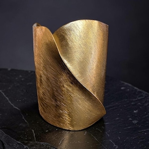 Può includere: Un bracciale a fascia larga con finitura in oro spazzolato. Il metallo presenta un distinto design diagonale sovrapposto che crea una silhouette curva e scultorea. La superficie presenta un motivo a grana fine e testurizzata su tutto il pezzo.