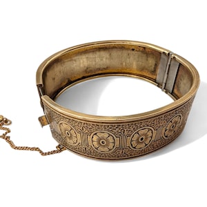May include: A gold-toned hinged bangle bracelet with an engraved floral pattern. The bracelet has a safety chain and clasp. The design features repeating floral motifs around the band. The bracelet is shown on a white background.