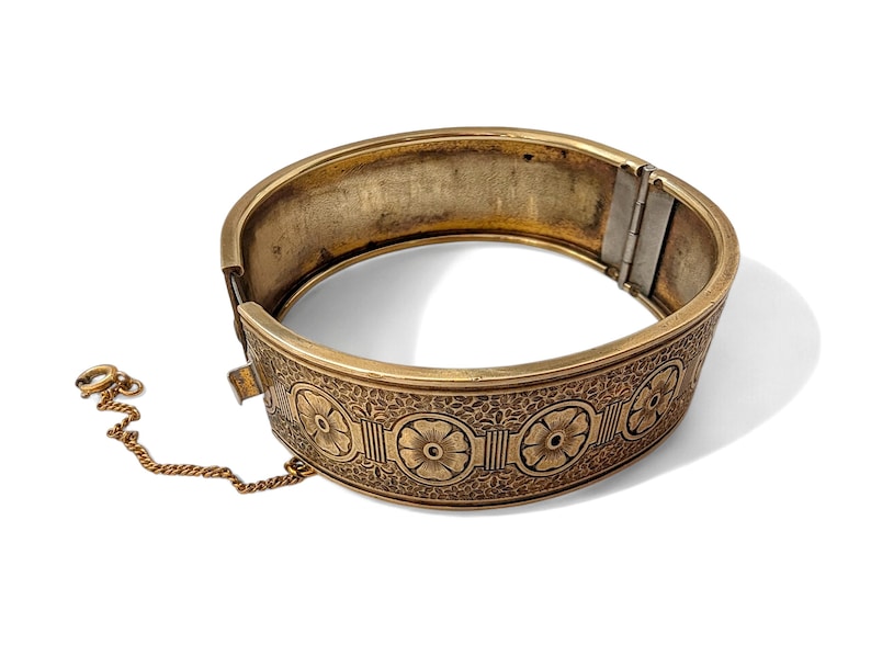 May include: A gold-toned hinged bangle bracelet with an engraved floral pattern. The bracelet has a safety chain and clasp. The design features repeating floral motifs around the band. The bracelet is shown on a white background.
