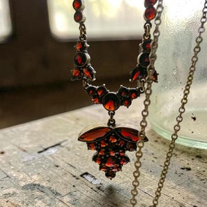 May include: A vintage necklace with a pendant featuring multiple red gemstones. The necklace has a delicate chain and an ornate design. The gemstones are set in a dark metal setting, creating a striking contrast.
