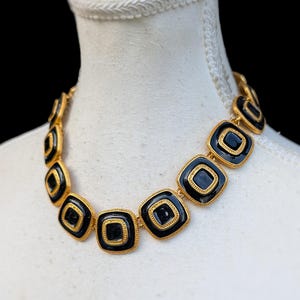 May include: A gold-tone metal necklace featuring a series of square links. Each link contains a glossy black enamel centre surrounded by a textured, beaded gold border. The necklace is displayed on a white mannequin against a black background.