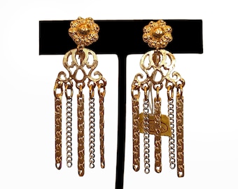 May include: A pair of gold-toned dangle earrings with a floral top design and multiple chain tassels. The earrings are displayed on a black stand against a white background. The earrings have a vintage aesthetic.