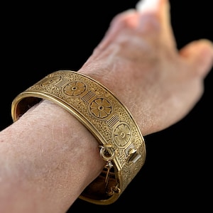 May include: A gold-colored hinged cuff bracelet with floral and geometric patterns. The bracelet is secured with a clasp. The design features repeating floral motifs and intricate detailing, creating a vintage aesthetic. The bracelet is approximately 2 inches wide.