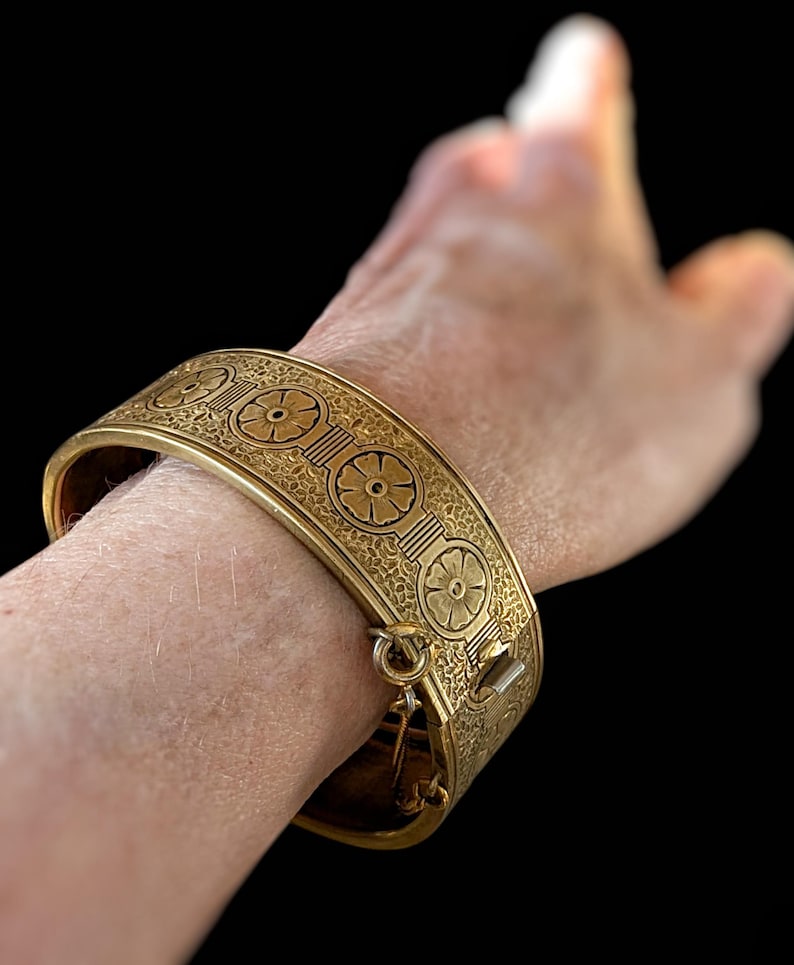 May include: A gold-colored hinged cuff bracelet with floral and geometric patterns. The bracelet is secured with a clasp. The design features repeating floral motifs and intricate detailing, creating a vintage aesthetic. The bracelet is approximately 2 inches wide.
