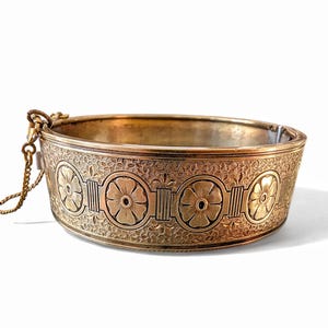 May include: A gold-toned metal bracelet with an engraved floral pattern. The bracelet features a series of flower designs and decorative elements. A small chain is attached to the bracelet. The bracelet is set against a white background.