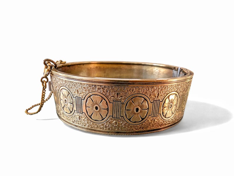 May include: A gold-toned metal bracelet with an engraved floral pattern. The bracelet features a series of flower designs and decorative elements. A small chain is attached to the bracelet. The bracelet is set against a white background.