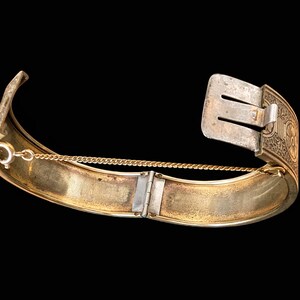 May include: A vintage gold-toned hinged bracelet with a silver-colored clasp. The bracelet features an ornate design on the clasp and a safety chain. The interior of the bracelet has a gold finish, and the exterior has a worn appearance.