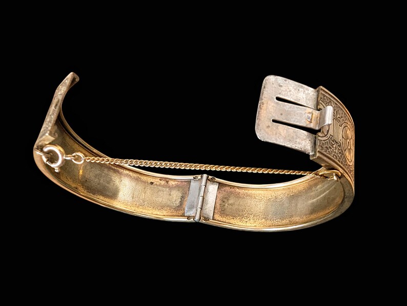 May include: A vintage gold-toned hinged bracelet with a silver-colored clasp. The bracelet features an ornate design on the clasp and a safety chain. The interior of the bracelet has a gold finish, and the exterior has a worn appearance.