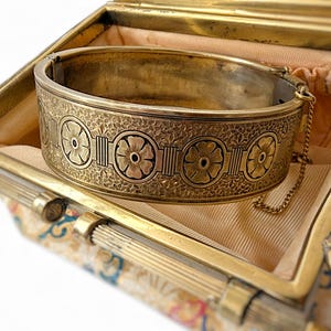 May include: A vintage gold-toned hinged bangle bracelet with floral and geometric designs. The bracelet is inside a matching gold-toned box with a patterned fabric lining. A delicate chain secures the bracelet.
