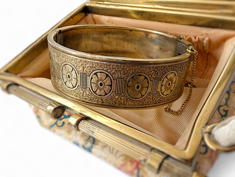 May include: A vintage gold-toned hinged bangle bracelet with floral and geometric designs. The bracelet is inside a matching gold-toned box with a patterned fabric lining. A delicate chain secures the bracelet.