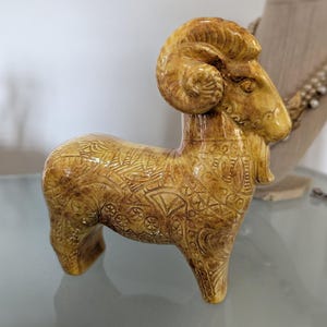 May include: A ceramic ram figurine in shades of tan and brown. The ram has large, curved horns and is decorated with intricate etched patterns. The figurine is standing on a reflective surface.