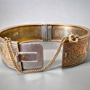 May include: An antique gold-tone hinged bangle bracelet. The bracelet features intricate floral and geometric patterns. A small gold chain is attached to the clasp for added security. The bracelet is approximately 2.5 inches in diameter.