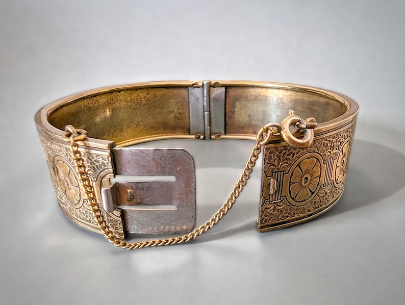 May include: An antique gold-tone hinged bangle bracelet. The bracelet features intricate floral and geometric patterns. A small gold chain is attached to the clasp for added security. The bracelet is approximately 2.5 inches in diameter.