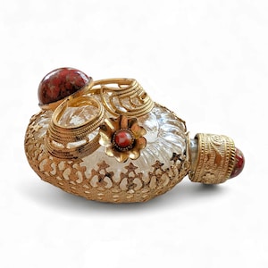 May include: An antique perfume bottle with a clear glass body and gold filigree detailing. The bottle has a red gemstone on the stopper and gold embellishments, displaying a vintage style. The bottle is likely made of glass.
