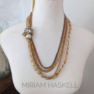 Puede incluir: Un collar de oro de múltiples hilos con un broche de perlas y piedras azules. El collar está formado por diferentes estilos de cadenas e incluye pequeñas cuentas doradas. El texto "MIRIAM HASKELL" es visible debajo del collar.