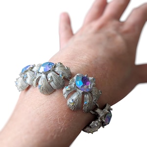 Può includere: Un braccialetto color argento con un design floreale con pietre preziose ovali e iridescenti. Il braccialetto ha una finitura in metallo spazzolato e strutturato ed è mostrato su un polso.