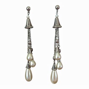 Può includere: Un paio di orecchini pendenti color argento con una parte superiore a forma di campana e una catena di piccole perline. Gli orecchini presentano tre perle finte a forma di pera, bianche sporche, appese alla catena.