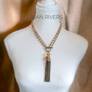 May include: A gold and brown beaded necklace with a tassel pendant. The necklace is on a white mannequin bust. The text "JOAN RIVERS" is visible above the necklace.