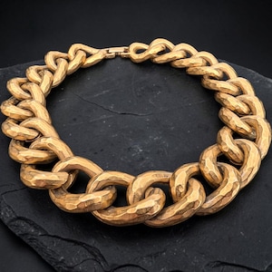 Puede incluir: Un collar de cadena gruesa en tono dorado con grandes eslabones ovalados entrelazados que presentan un acabado de superficie martillada y texturizada. El collar está diseñado como un bucle circular continuo con un cierre visible en el centro.