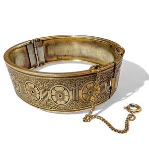 May include: A vintage gold-toned bracelet with a floral pattern and a safety chain. The hinged bracelet features a detailed design of flowers and foliage. The clasp is open, revealing the interior of the bracelet.