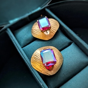May include: A pair of gold-toned clip-on earrings with large, rectangular, iridescent, ruby-colored gemstones. The earrings are set on textured, circular bases and are displayed in a teal jewelry box.