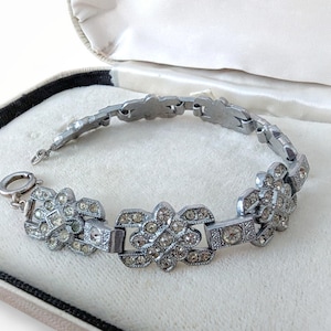May include: A silver-toned bracelet with a floral design, set with clear rhinestones. The bracelet is displayed in an open, cream-colored jewelry box with a black trim. The clasp is a simple ring and bar closure.