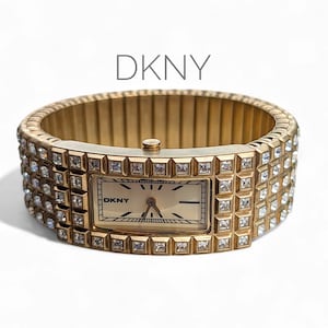 May include: A gold-coloured DKNY watch with a rectangular face and a band embellished with square-cut crystals. The watch face is cream with black hands and markers. The band is a stretch style.