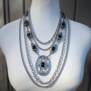 Può includere: Collana multistrato color argento con pietre nere di forma ovale e un ciondolo ornato. La collana è esposta su un manichino bianco, mettendo in risalto il suo design a strati e i dettagli intricati. Il ciondolo è lungo circa 5 cm.