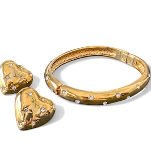 Op de afbeelding: Een goudkleurige sieradenset met een scharnierende armband en hartvormige oorbellen. De armband en oorbellen zijn versierd met heldere, sprankelende edelstenen. De oorbellen hebben een sterrenpatroon. De sieraden liggen op een witte achtergrond.