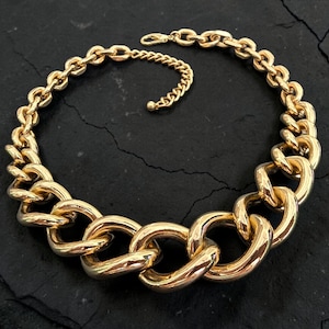 May include: A gold-toned chunky chain necklace featuring large, polished interlocking oval links that graduate in size towards the centre. The necklace includes a shorter, thinner chain segment with a small clasp for adjustable length and secure closure.